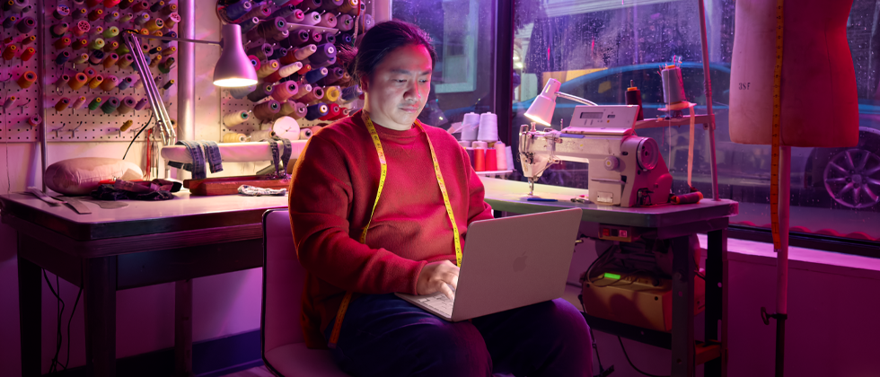 Iemand met MacBook Neo in de kleur blos op schoot zonder netsnoer, in een werkruimte met een naaimachine en bijbehorende materialen, waaronder garen en een paspop