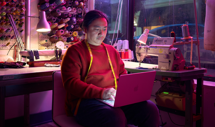 Iemand met MacBook Neo in de kleur blos op schoot zonder netsnoer, in een werkruimte met een naaimachine en bijbehorende materialen, waaronder garen en een paspop