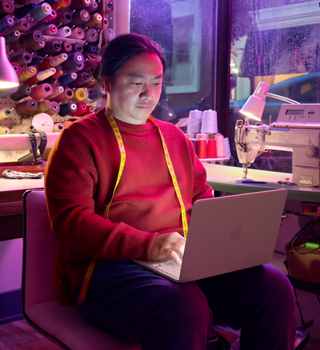 Iemand met MacBook Neo in de kleur blos op schoot zonder netsnoer, in een werkruimte met een naaimachine en bijbehorende materialen, waaronder garen en een paspop