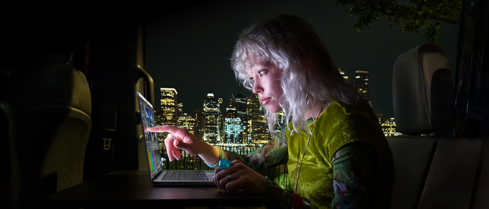 Iemand zit op de achterbank van een bus, achter de open passagiersdeur is de skyline van een stad in het donker te zien, MacBook Pro met PROLED XDR-display op een werktafel, de persoon wordt verlicht door het scherm terwijl die de Touch-feature gebruikt om een video te bewerken in DaVinci Resolve Studio