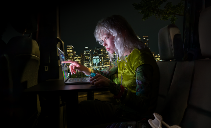 Iemand zit op de achterbank van een bus, achter de open passagiersdeur is de skyline van een stad in het donker te zien, MacBook Pro met PROLED XDR-display op een werktafel, de persoon wordt verlicht door het scherm terwijl die de Touch-feature gebruikt om een video te bewerken in DaVinci Resolve Studio