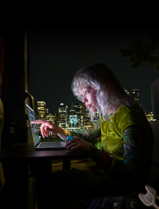 Iemand zit op de achterbank van een bus, achter de open passagiersdeur is de skyline van een stad in het donker te zien, MacBook Pro met PROLED XDR-display op een werktafel, de persoon wordt verlicht door het scherm terwijl die de Touch-feature gebruikt om een video te bewerken in DaVinci Resolve Studio