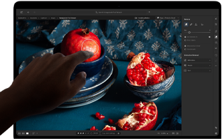 MacBook Pro met PROLED XDR-display met een stilleven van granaatappels met blauw keramiek en textiel, een hand reikt naar het scherm om de creatie aan te raken
