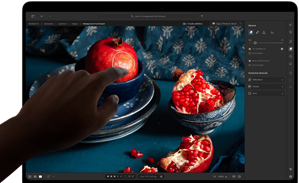 MacBook Pro met PROLED XDR-display met een stilleven van granaatappels met blauw keramiek en textiel, een hand reikt naar het scherm om de creatie aan te raken