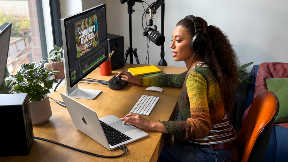 Een vrouw werkt thuis aan haar bureau met een MacBook Pro die is aangesloten op twee externe displays. Ze draagt een koptelefoon en praat in een microfoon