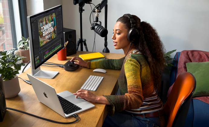 Een vrouw werkt thuis aan haar bureau met een MacBook Pro die is aangesloten op twee externe displays. Ze draagt een koptelefoon en praat in een microfoon